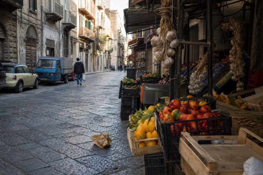 A busy Palermo street market with crates of citrus and hanging strings of garlic, morning light