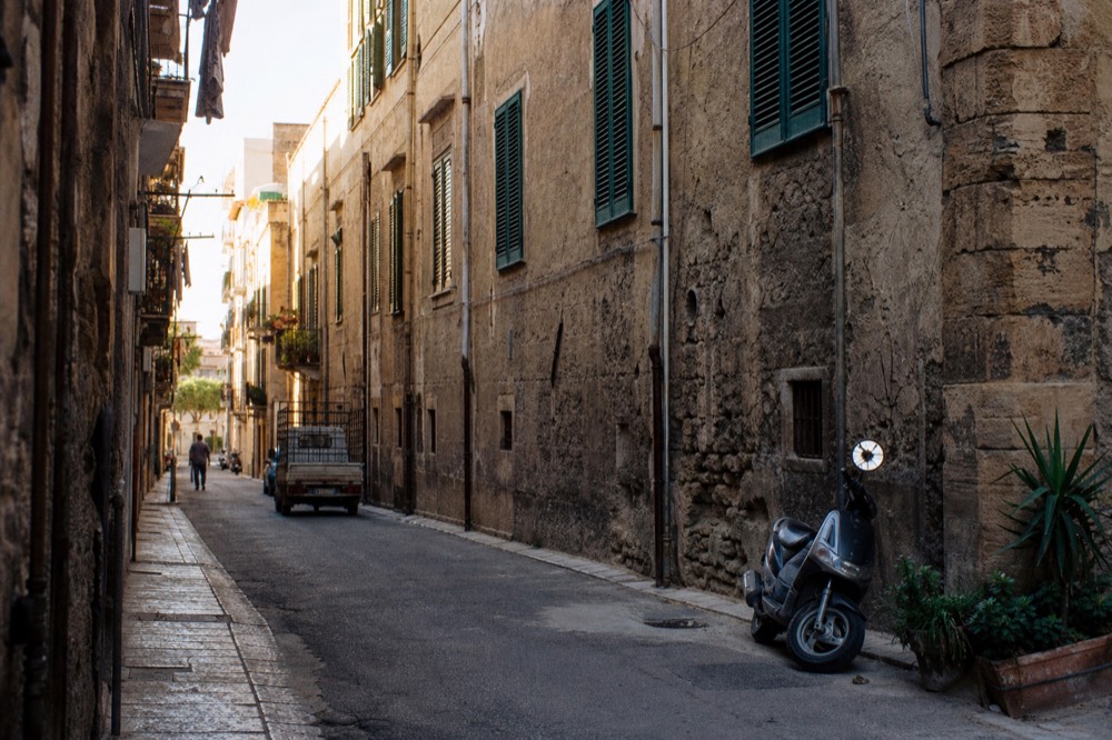 A narrow Palermo side street with faded shutters and stone buildings rising on both sides