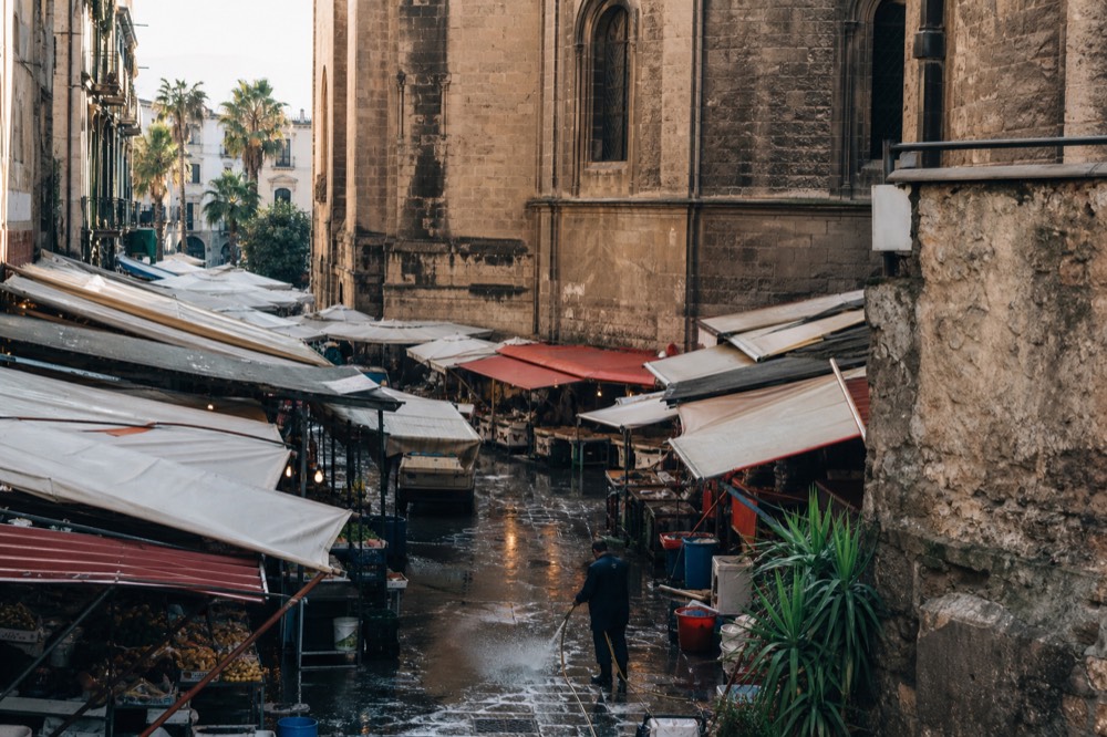 A crowded Palermo market street with stalls of produce and hanging canvas awnings