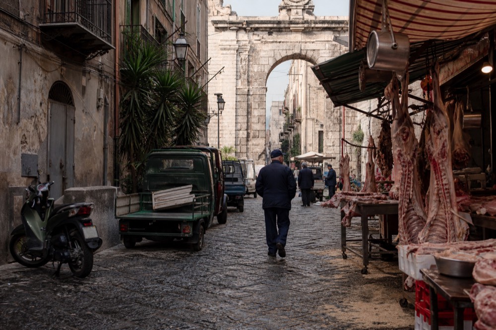 Butcher and produce stalls in a narrow Palermo market lane with signs overhead