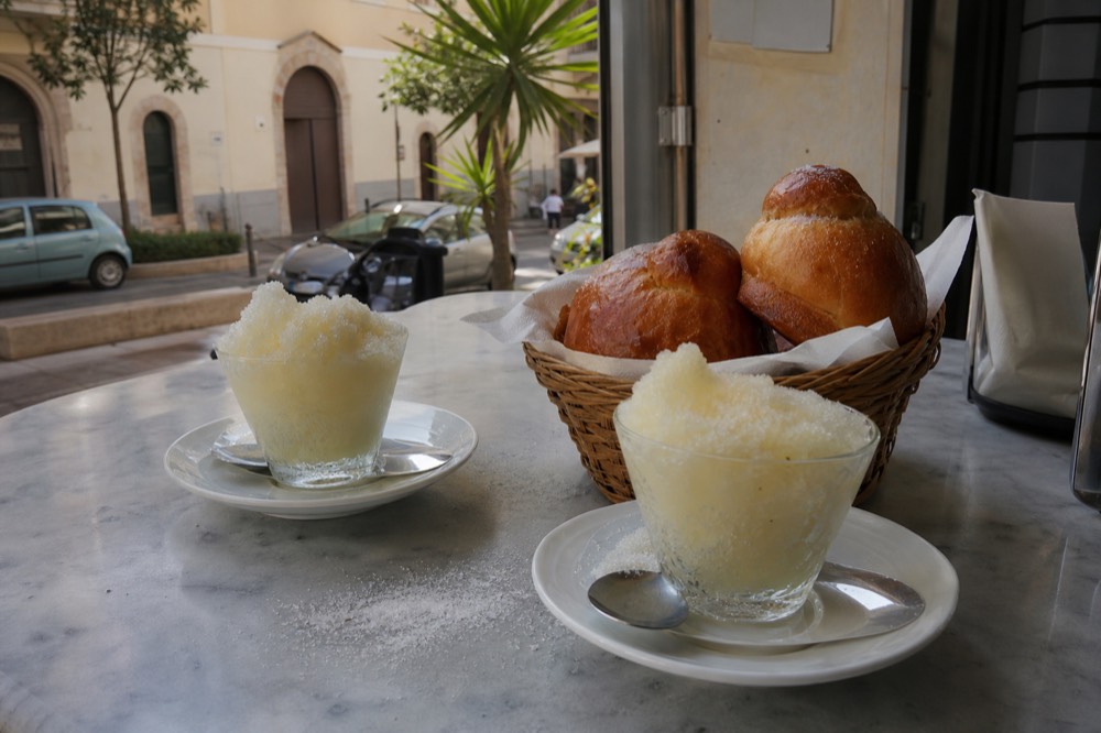 Two glasses of almond granita on a cafe counter beside a basket of sweet brioches