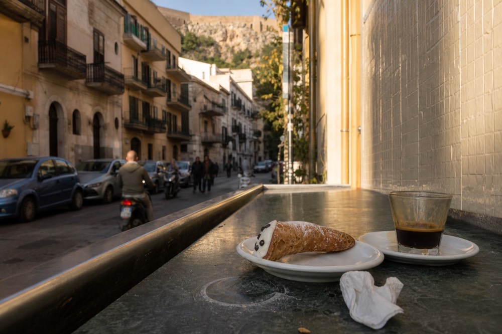 A cannolo filled with ricotta cream on a small plate beside a cup of espresso
