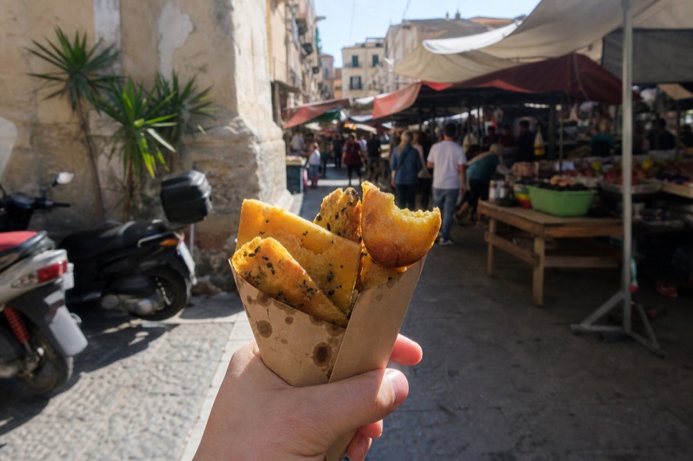 A paper cone of fried panelle and crocche held above a Palermo market street