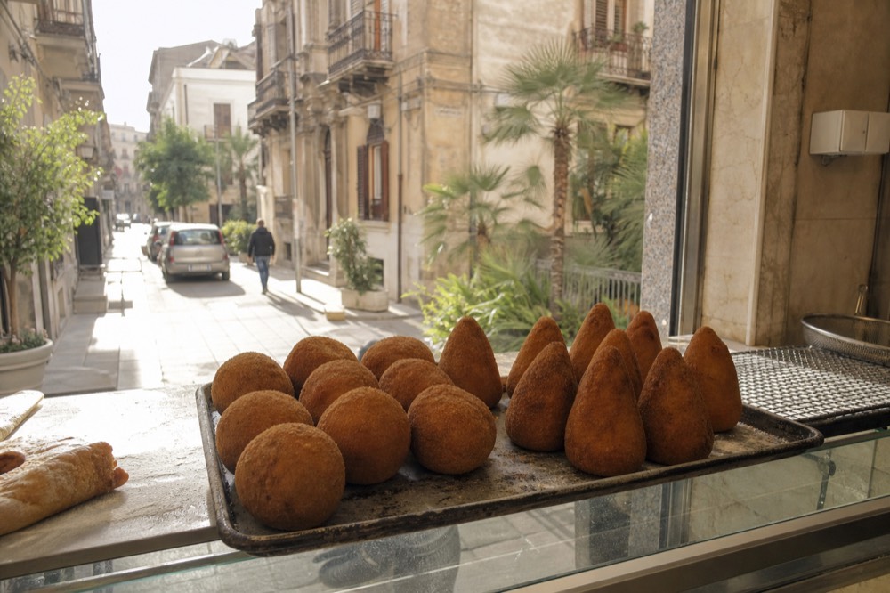 Round Sicilian arancine beside cone shaped arancini on a bakery counter