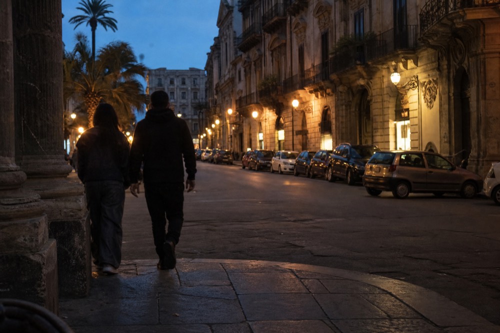 An evening street in Palermo with lit shopfronts and stone buildings along a straight road