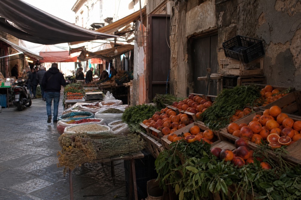 Market stalls with crates of oranges, herbs and spices under canvas awnings in a Palermo street market