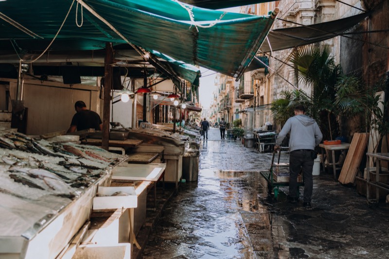 Fish on ice and vegetables under market awnings in a Palermo market street.