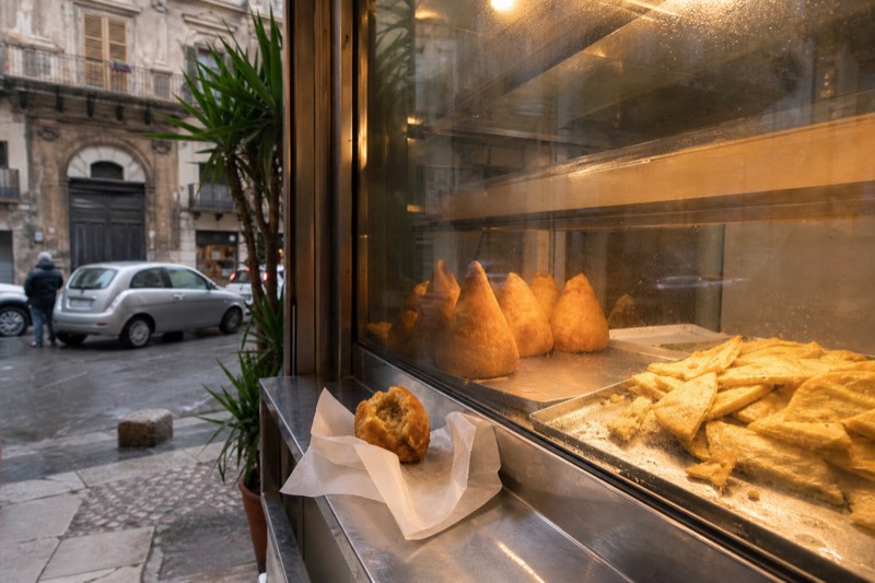 Arancine and panelle stacked in a glass case at a Palermo street food counter.