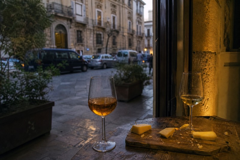 Glasses of Sicilian wine beside local cheeses on a board in a small tasting bar.