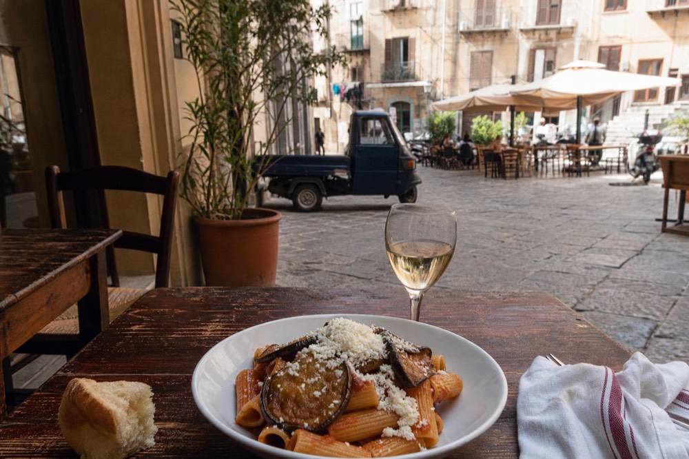 A bowl of pasta with eggplant and ricotta beside a glass of white wine on a wooden table