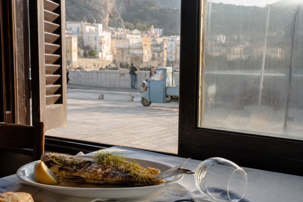 A plate of grilled fish with lemon and herbs on a table beside a window