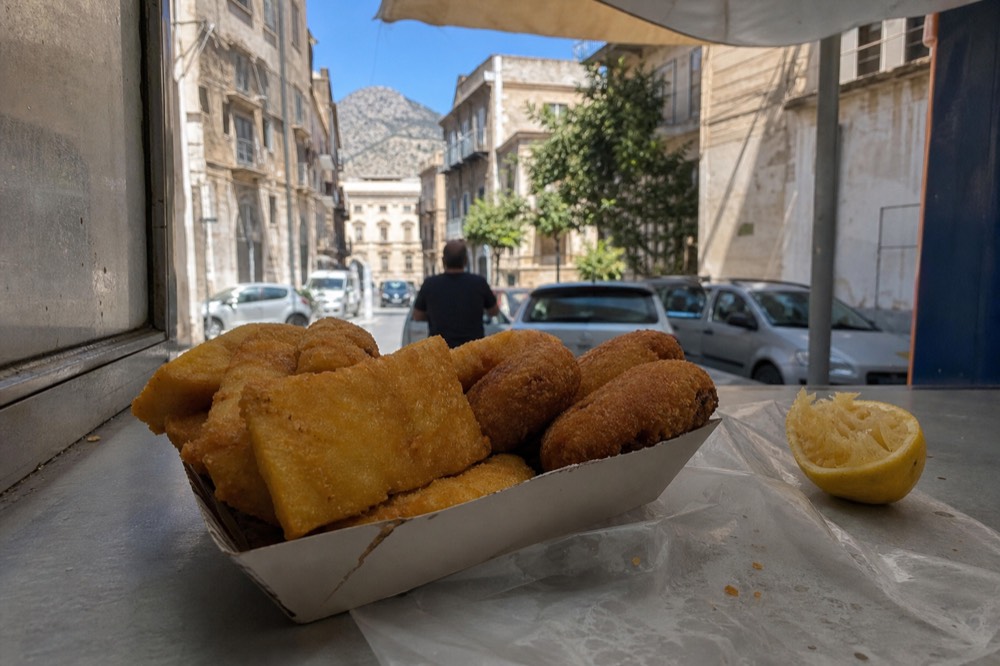 Fried chickpea fritters and potato croquettes stacked in a paper tray at a market counter