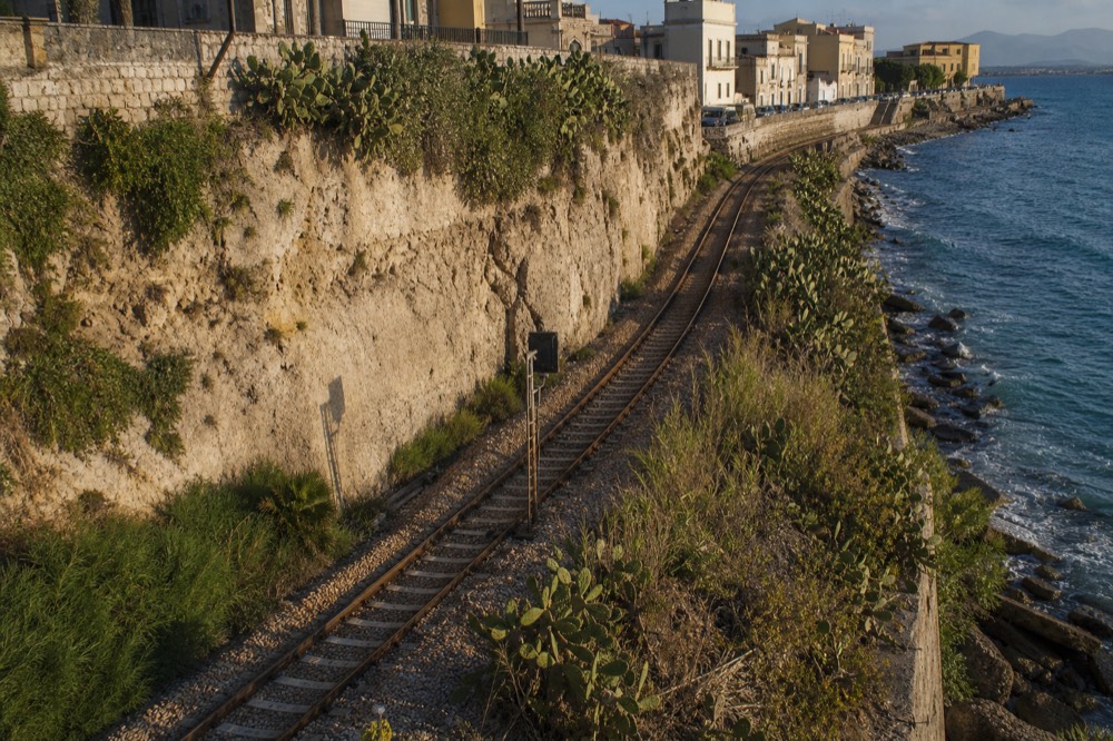 A single-track rail line running along a rocky Sicilian coastline