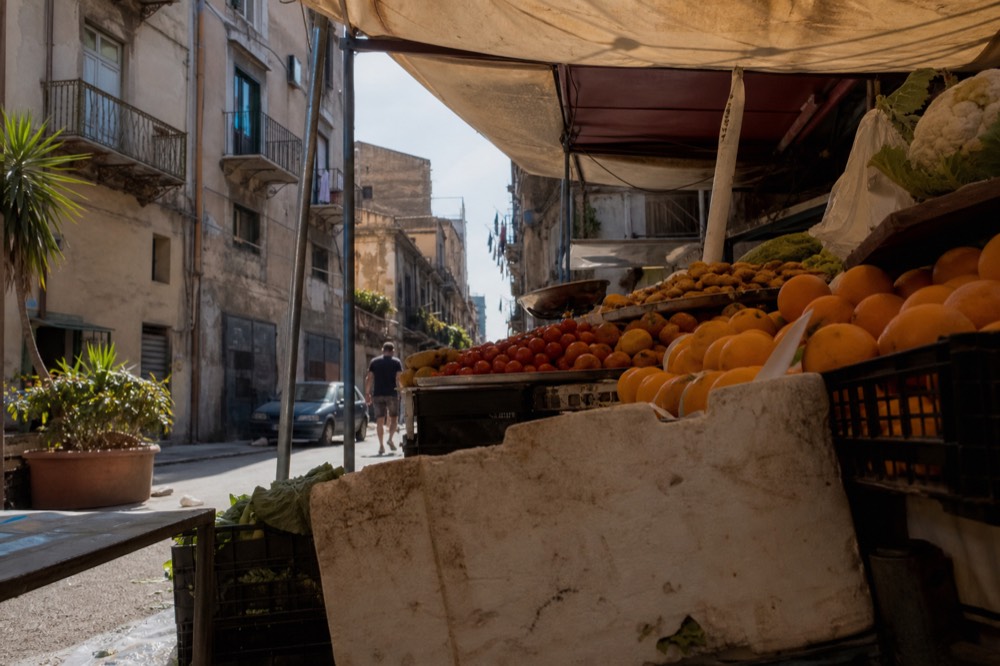 A market stall counter stacked with fresh produce and fried snacks under a canvas awning