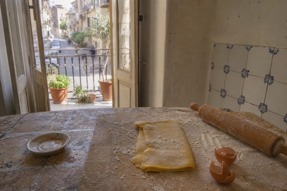 A rustic kitchen counter with rolled fresh pasta dough, flour and a wooden board