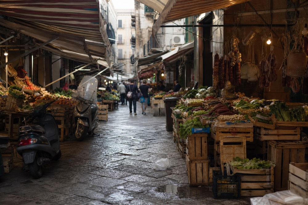 A crowded Palermo market lane with striped awnings and vegetable stalls