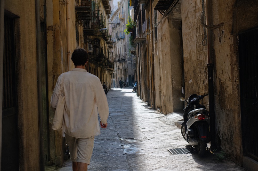 A visitor walking a narrow Palermo street between tall ochre buildings