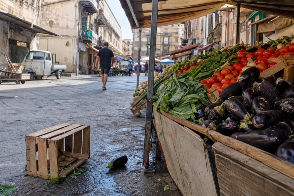 A market stall piled with aubergines, tomatoes and leafy greens under a canvas awning