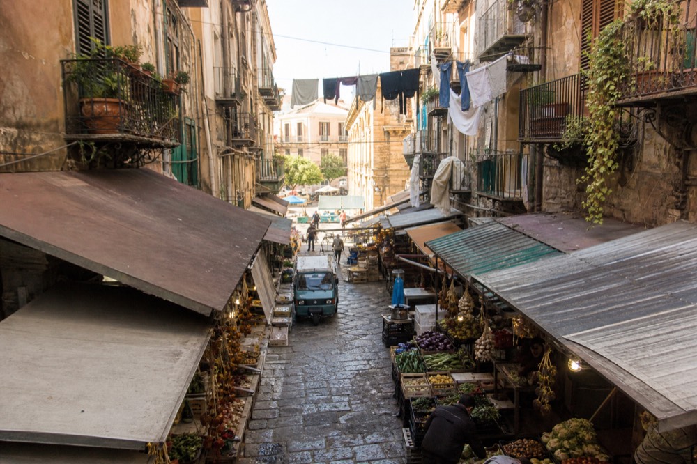 A busy Palermo market alley in the morning with hanging produce and awnings