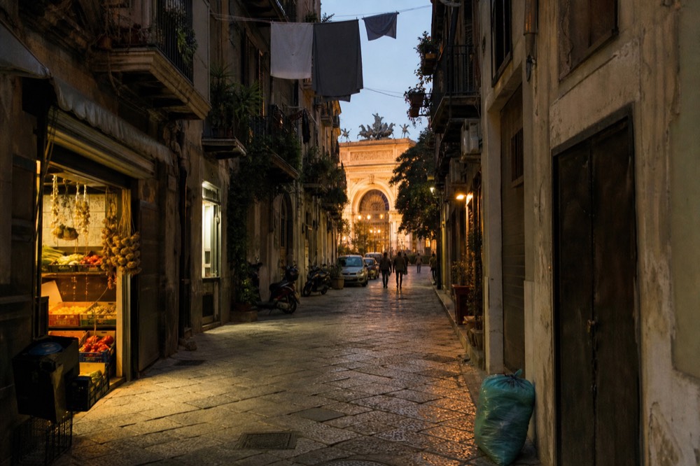 A narrow Palermo street with laundry overhead and a small grocery shop open in the evening