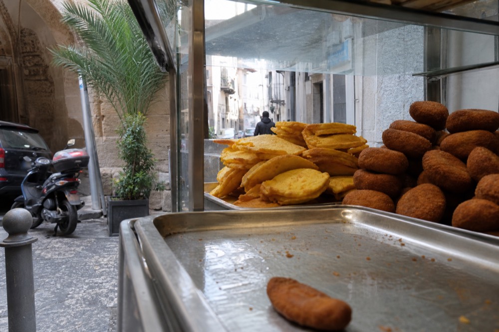 A glass display case of fried chickpea fritters and potato croquettes in a Palermo shop