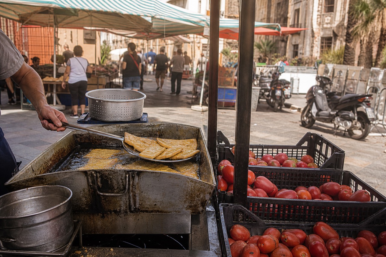 Panelle frying at a street cart beside packed produce stalls in central Palermo.