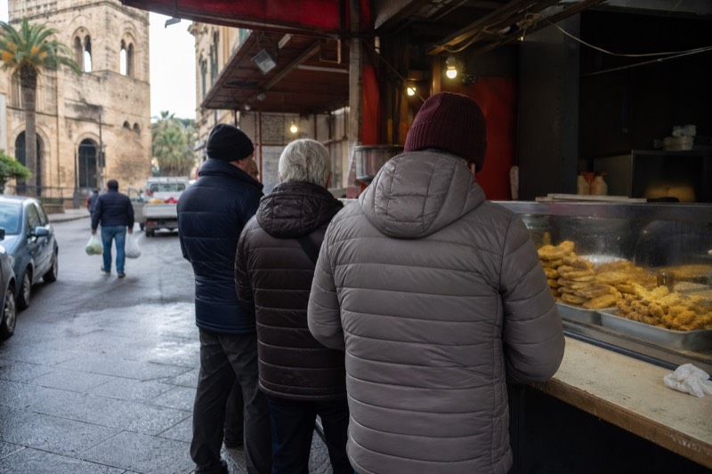 A queue at a Palermo street food counter, trays of fried snacks stacked behind the glass.